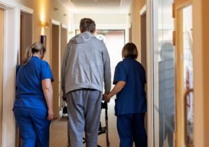 One Hospice patient is being accompanied by two nurses walking down a brightly lit corridor